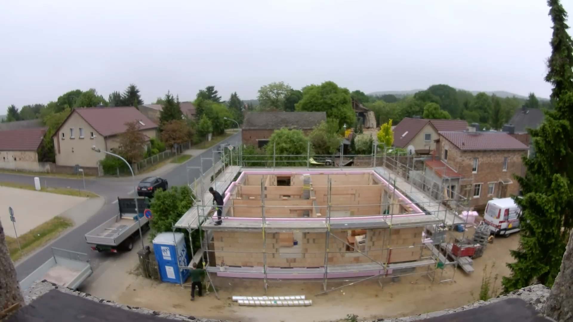 Mauerwerk Hausbau: Das Bild zeigt ein Haus von oben. Um das Haus ist ein zweistöckiges Gerüst auf dem ein Mann steht. Beim Haus steht nur das Mauerwerk