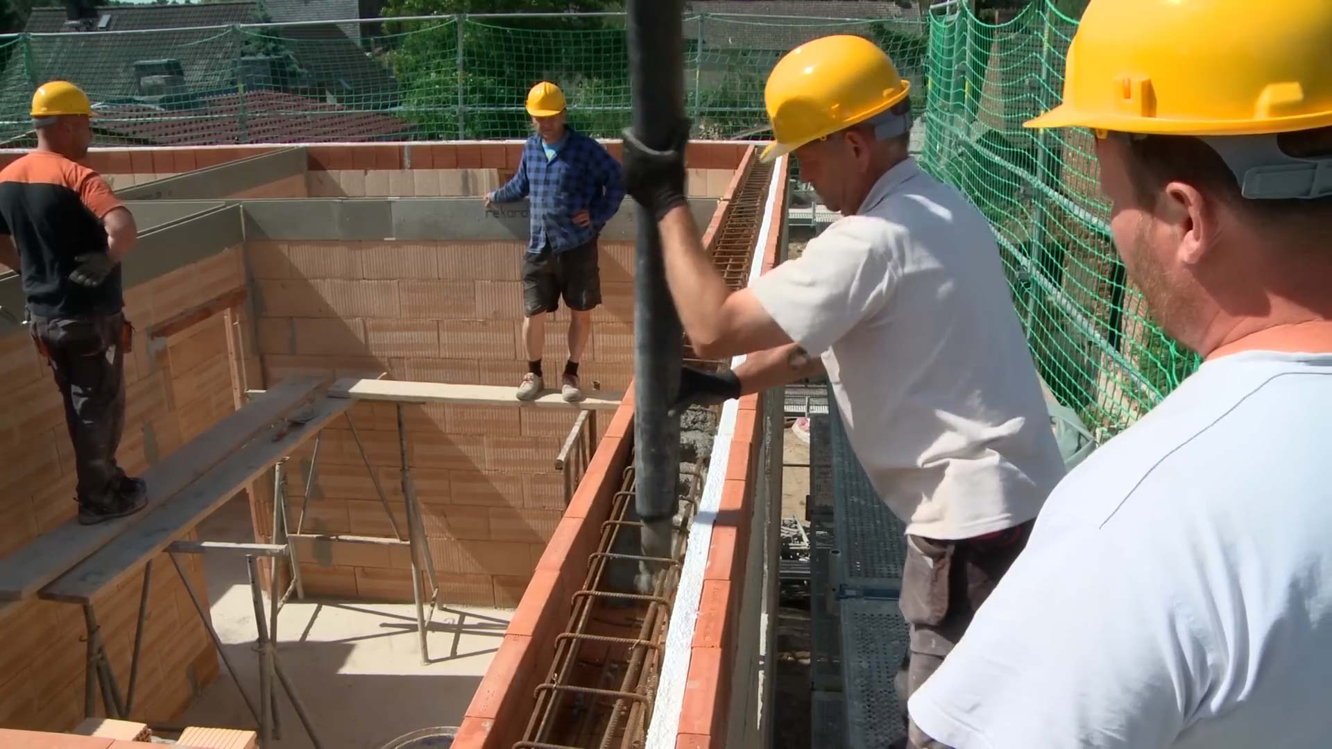 Mauerwerk Hausbau: Ein Mann füllt Beton mithilfe eines Rohres in den Ringanker eines Hauses. Im Hintergrund sind weitere Männer auf einem Gerüst um das Haus herum zusehen