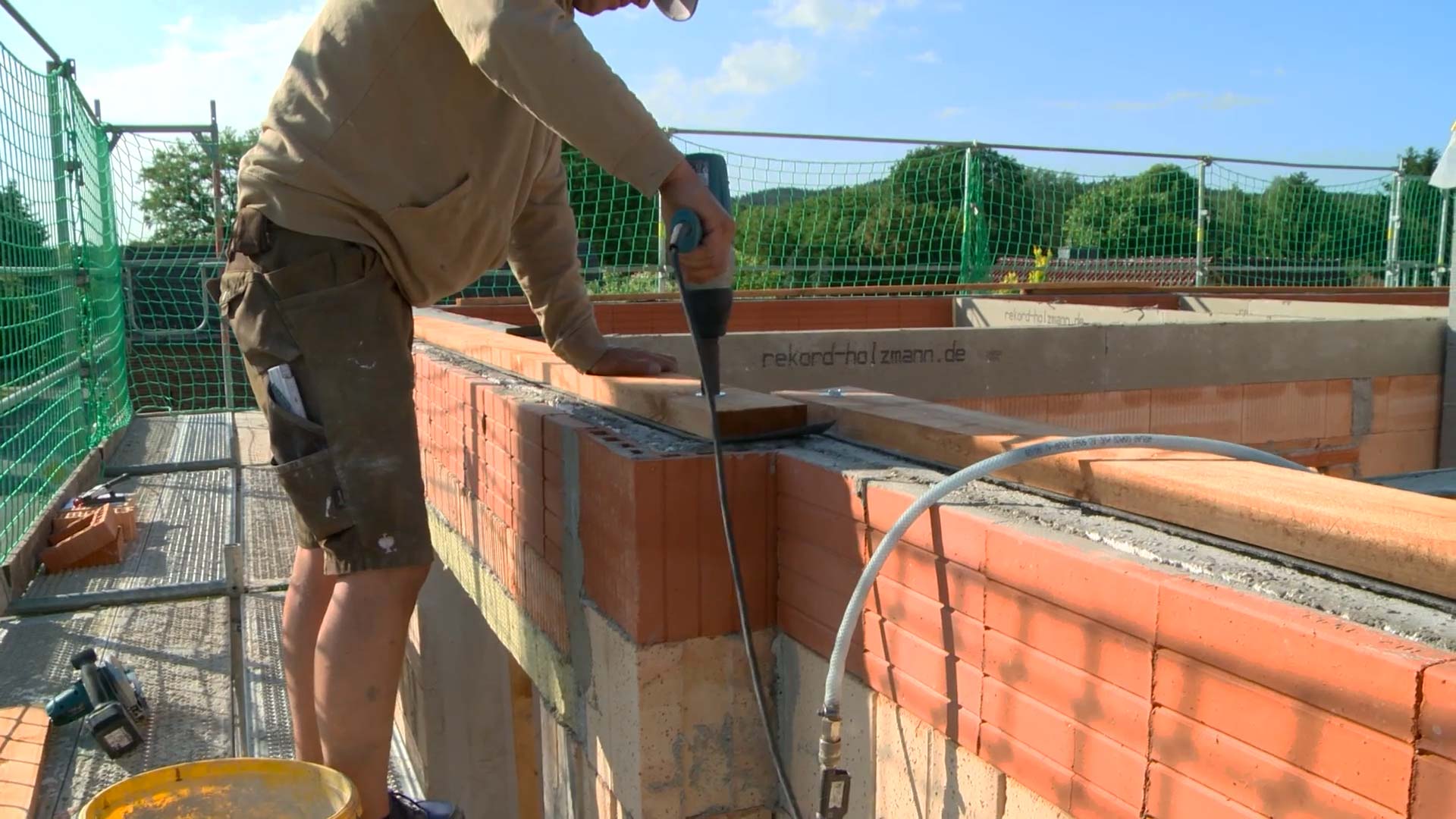 Dachstuhl bauen: Ein Mann bohrt einen Nagel in die Fußpfette des neuen Dachstuhls. Er steht auf einem Gerüst neben einem Rohbau.