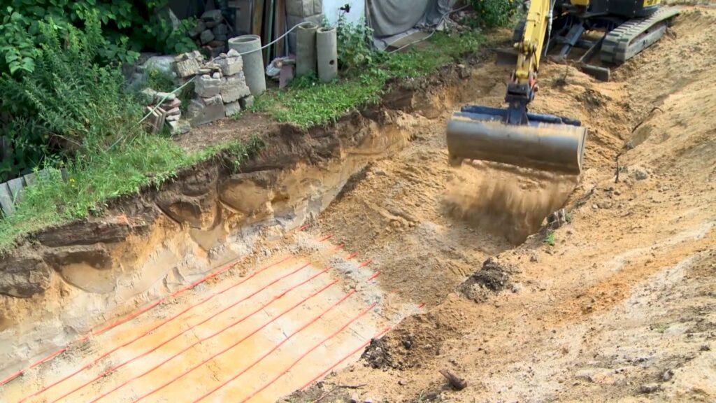 Rohinstallation Sanitär und Heizung: Ein Bagger verteilt Sand und Erde über die verlegten Wärmekollektoren-Rohre