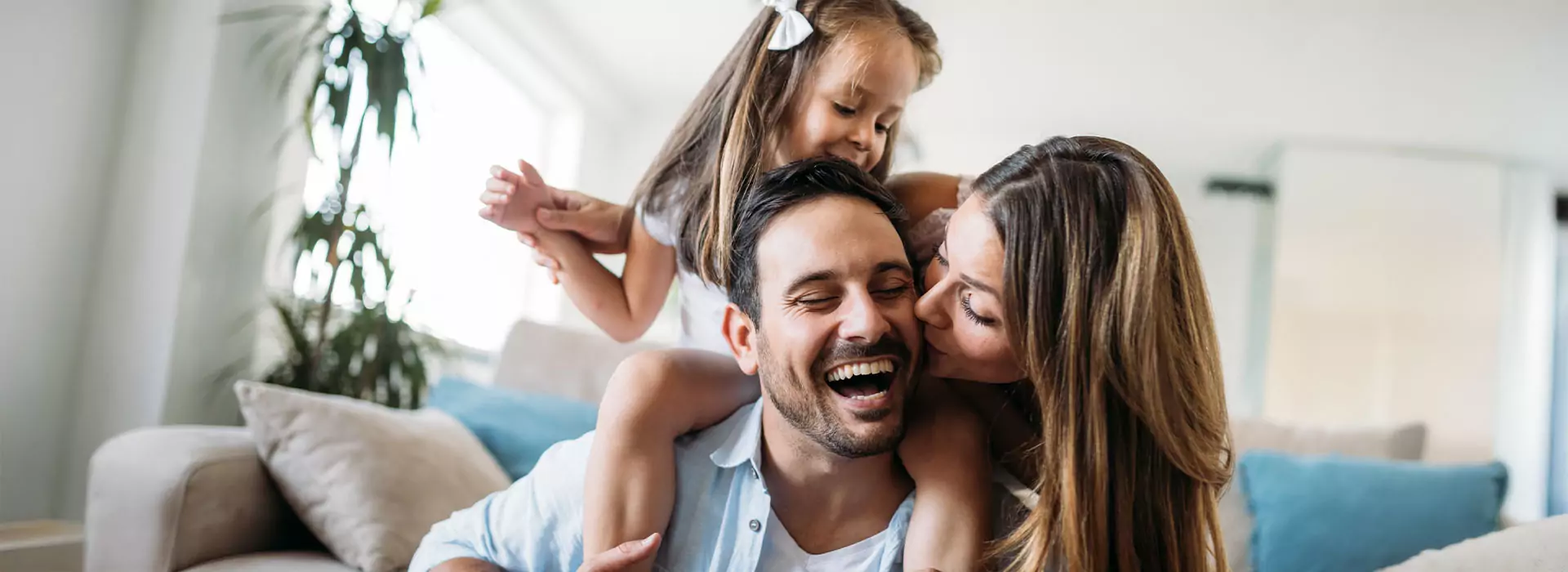 Das Bild zeigt eine lachende Familie mit einem Kind. Das Kind sitzt auf den Schultern des Mannes. Die Frau steht daneben.